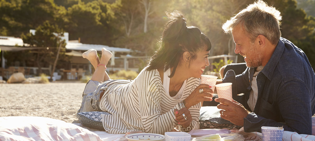 couple lounging on a blanket on a beach while toasting with cups and smiling at each other