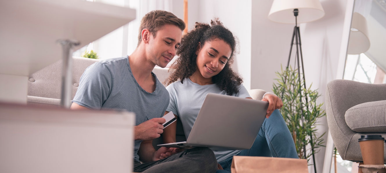 a young couple sits on the floor together in front of a laptop with a credit card in hand