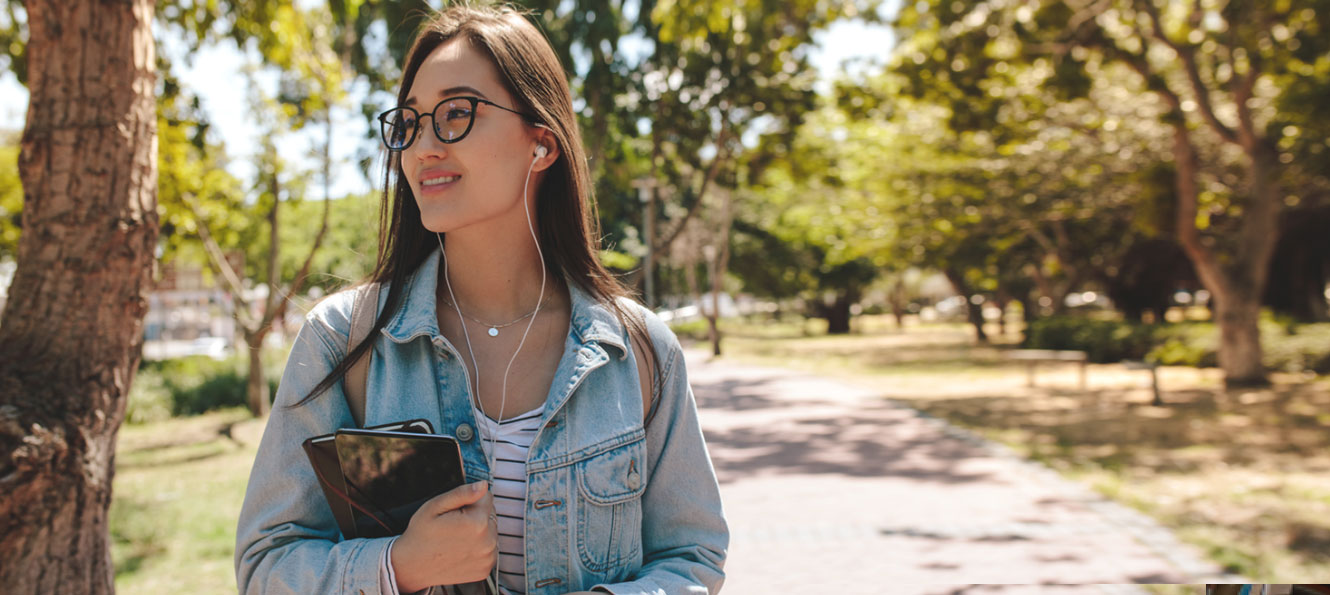 student wearing headphones while walking along a tree-lined campus path on a sunny day