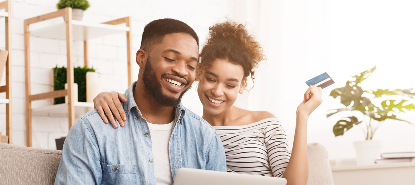 couple sitting on a couch together with a laptop - woman is holding a credit card in one hand