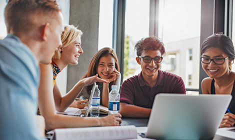 five students sitting around a table talking and looking at a computer together