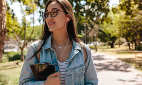 student wearing headphones while walking along a tree-lined campus path on a sunny day