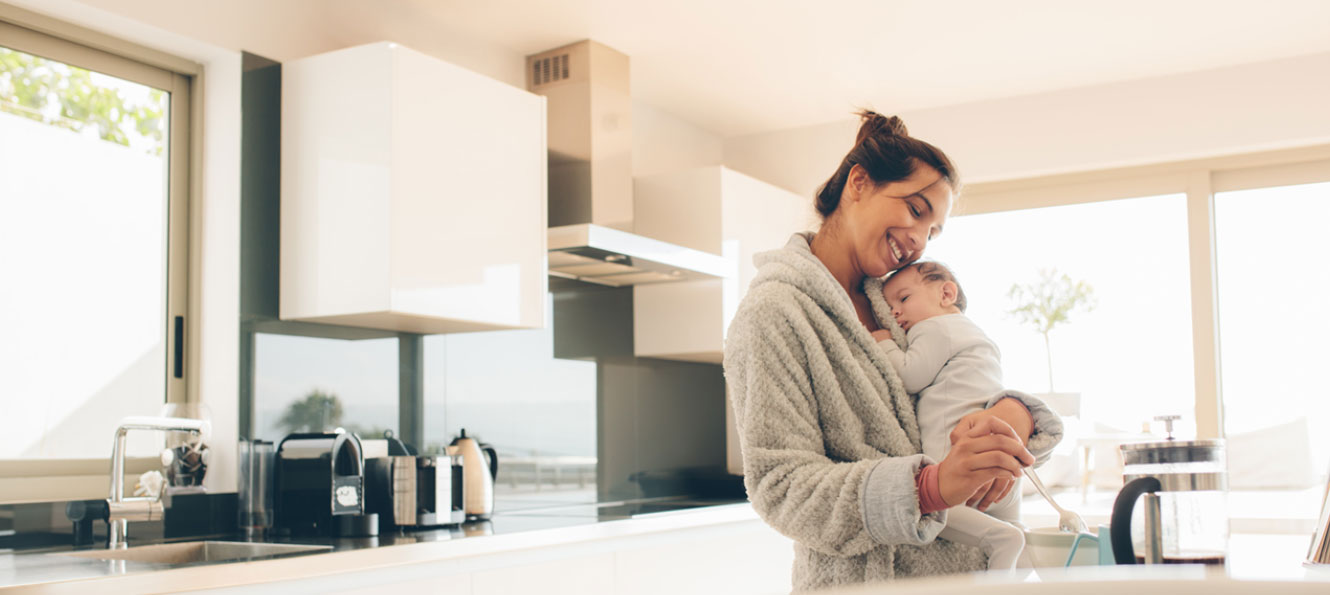 mother holding baby while she makes herself coffee in the morning