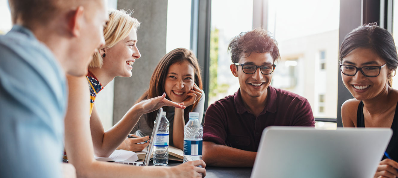 five students sitting around a table talking and looking at a computer together