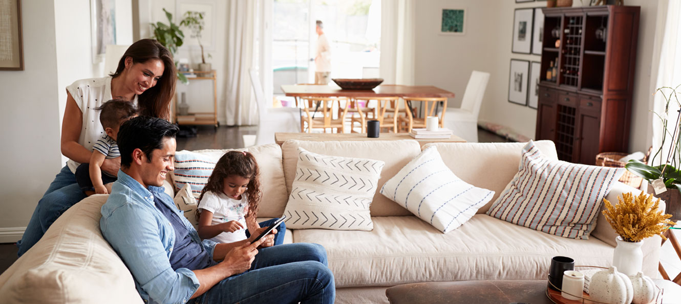 family of four sitting on a couch at home, looking at the tablet device the dad is holding